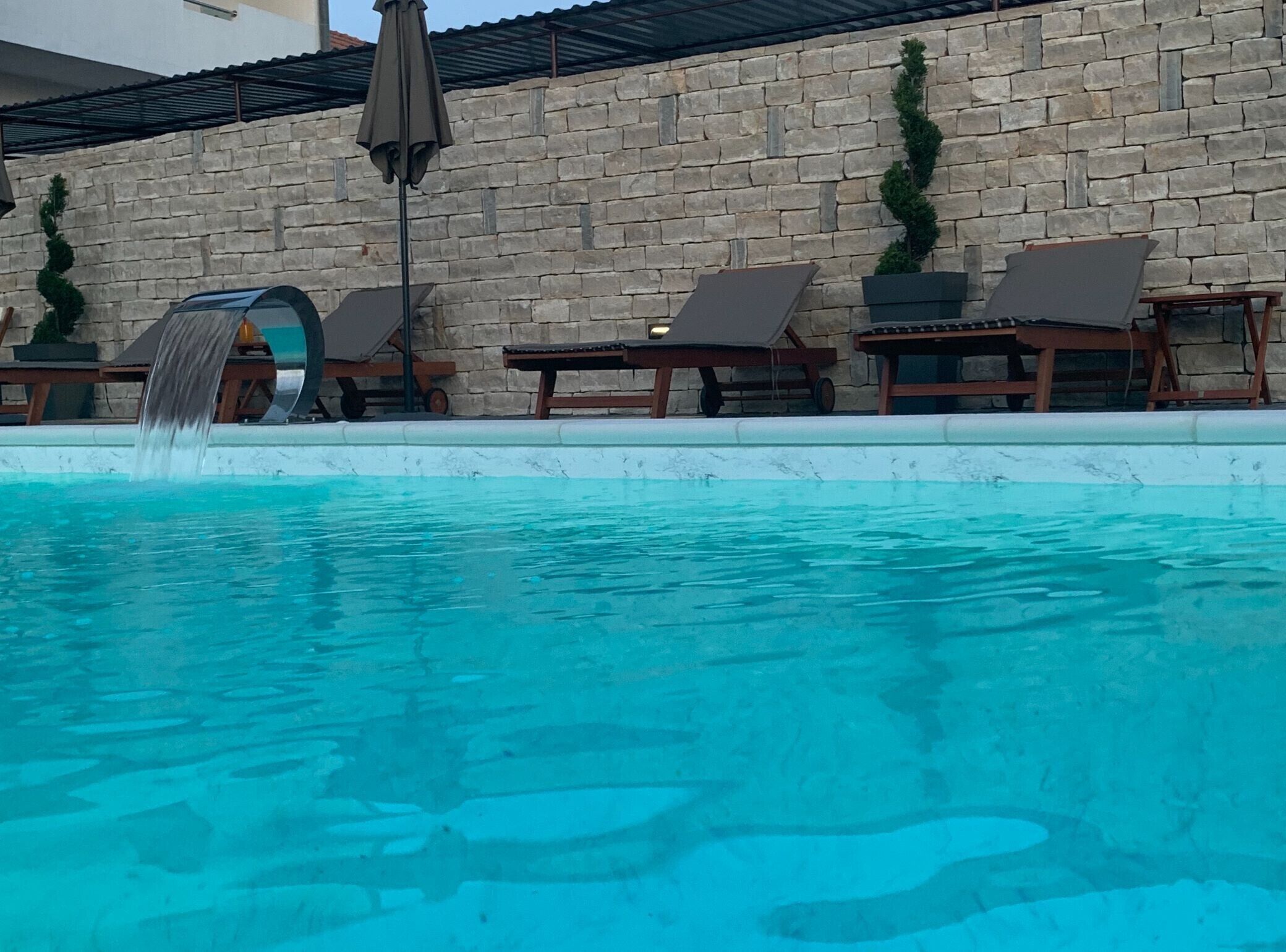 Una piscina con cascada, rodeada de sillas de descanso y un muro de piedra.