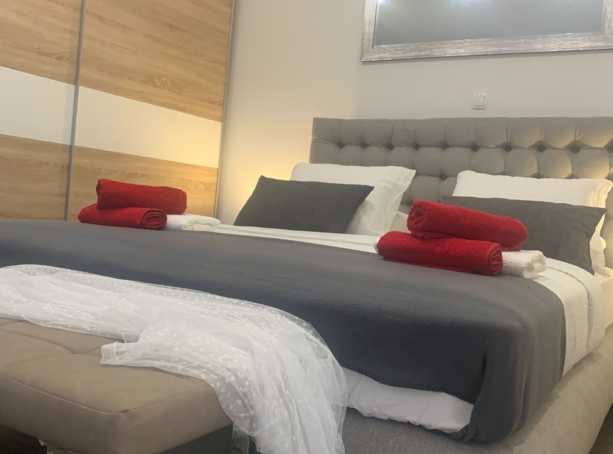 Ein ordentlich bezogenes Bett mit grauem Kopfteil, weißer und grauer Bettwäsche und roten Handtüchern.