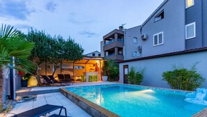 A rectangular swimming pool with a wooden deck, surrounded by lounge chairs and a small bar area. - Three Bedroom Apartment with Private Pool (Zadar)