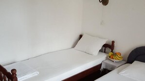 A single bed with a wooden frame and white bedding, a small table with a fruit bowl, and a wall-mounted light fixture.