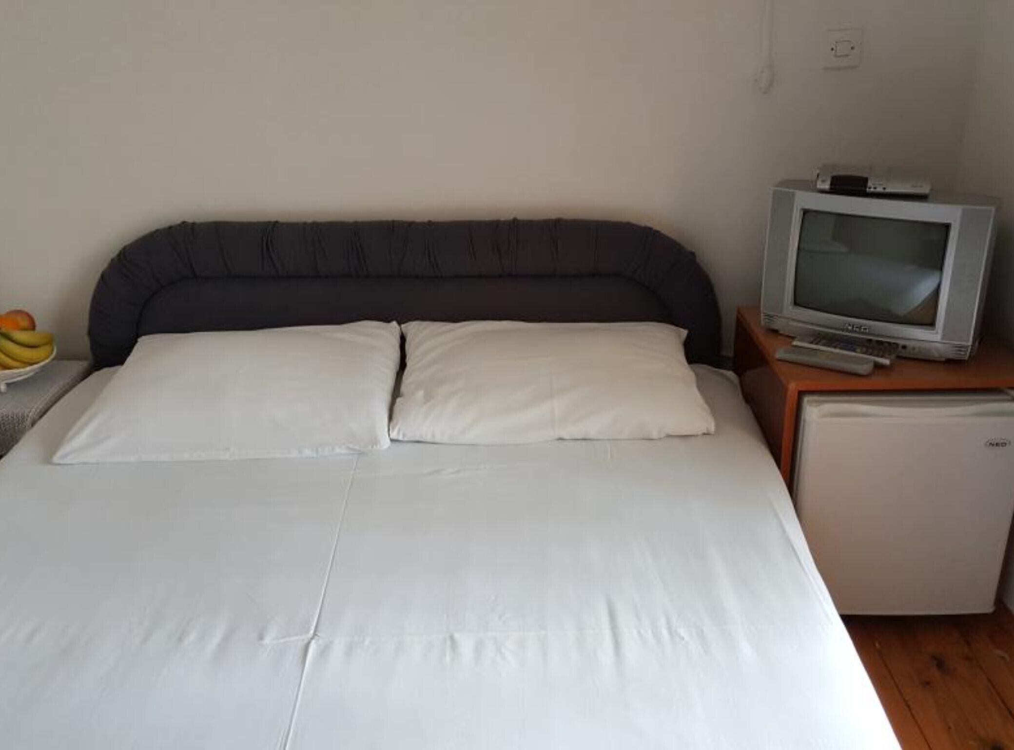 Een bed met witte lakens, een kleine televisie op een houten nachtkastje en een koelkast.
