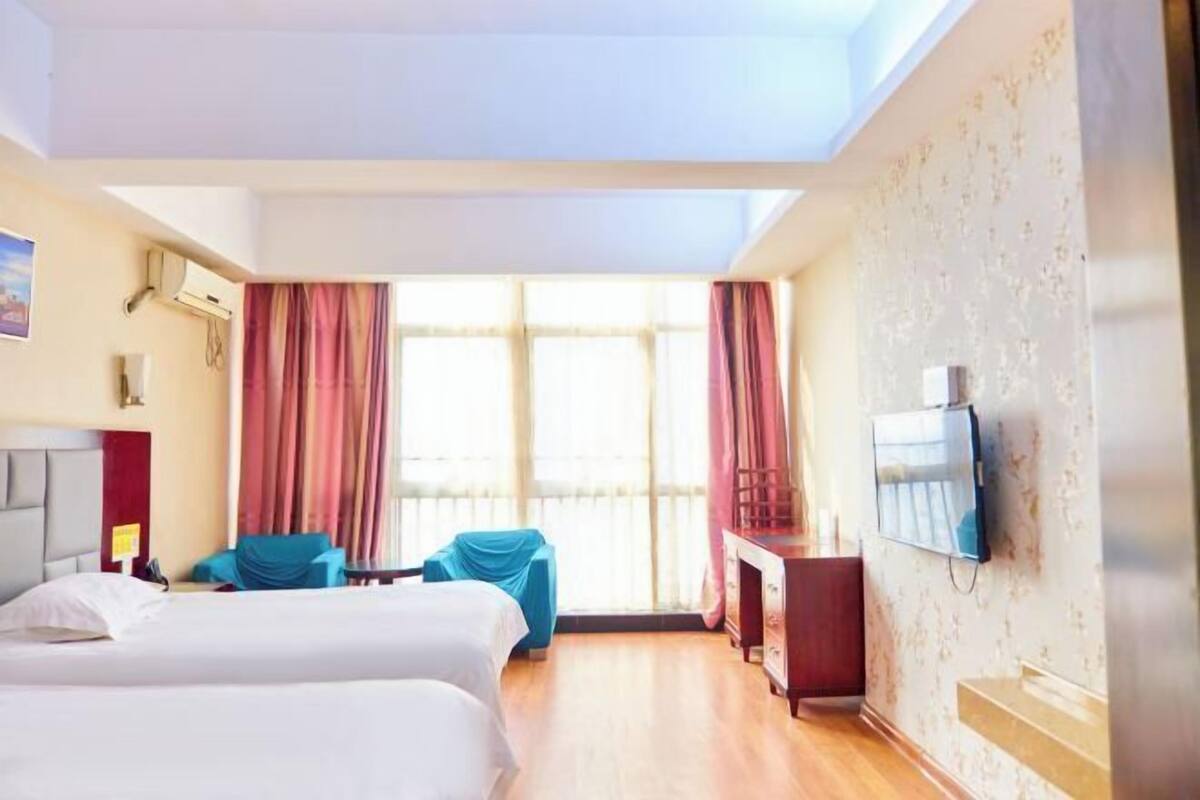 A hotel room with two beds, a desk, a TV, and large windows with curtains.