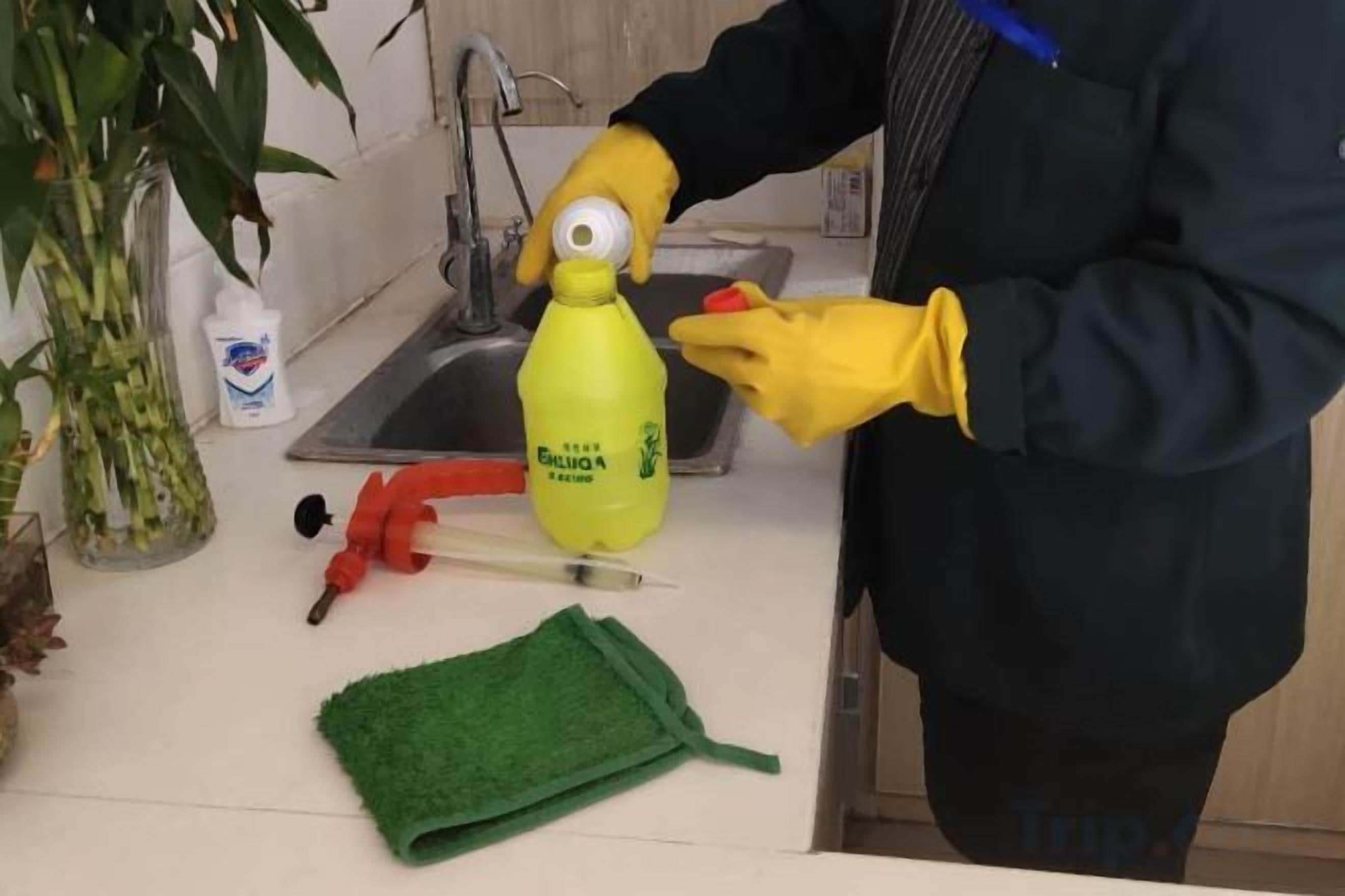Une personne portant des gants jaunes manipule une bouteille jaune munie d’une étiquette, et se tient dans une cuisine équipée d’un évier, d’un robinet et d’un torchon vert posé sur le comptoir.