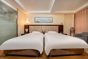 Habitación de hotel con dos camas individuales, un ventanal grande, un cuadro con una escena de playa y un sofá.