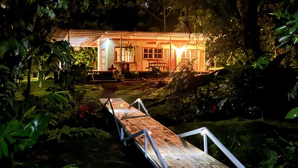 A wooden bridge leading to a house with a tiled roof, surrounded by lush greenery.