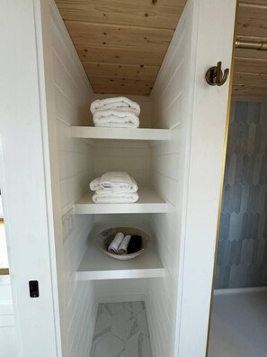 Ein schmaler begehbarer Kleiderschrank mit weiß gefliesten Wänden und Holzboden. Er beherbergt drei Regale mit gefalteten weißen Handtüchern und eine kleine Schale am unteren Rand.