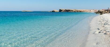A beach with clear turquoise water and white sand, with rocky formations in the background.