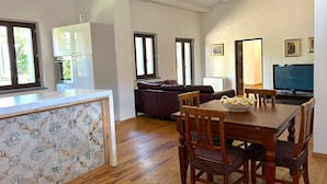 Una cocina moderna con armarios blancos, un comedor con mesa y sillas de madera, y una sala de estar con sofá y televisor.