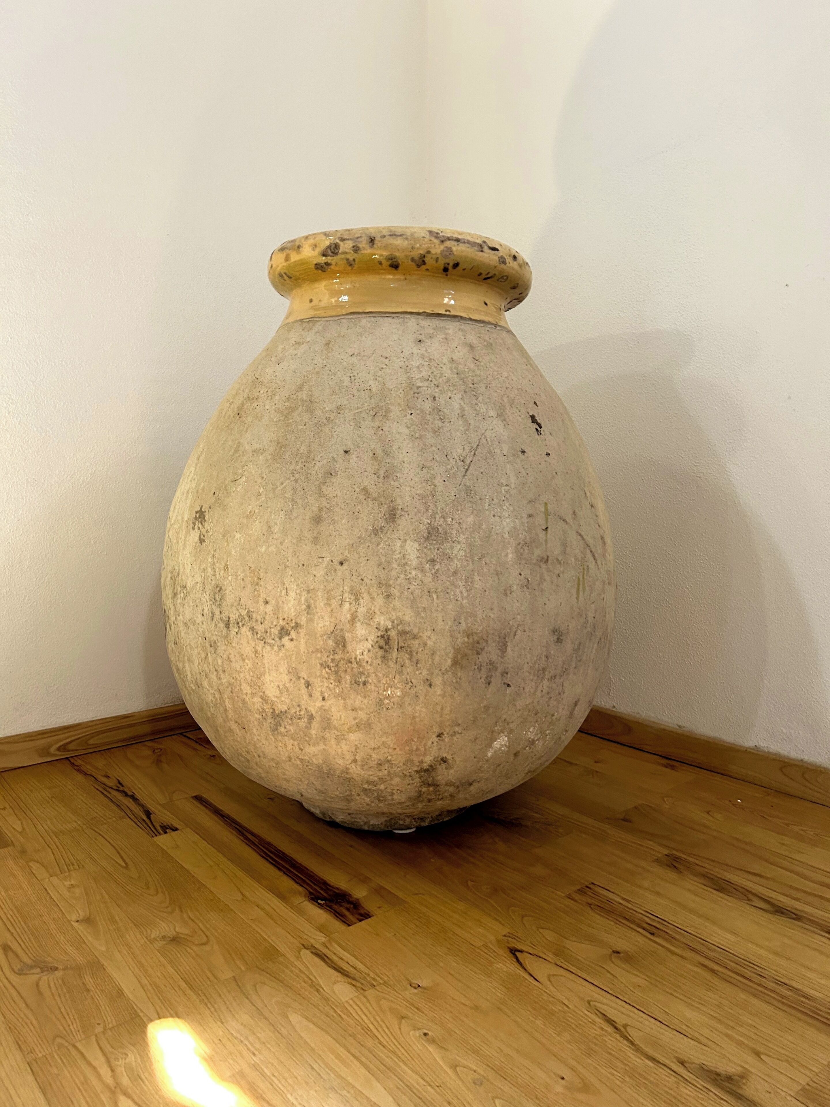 A large, round, beige ceramic vase with a narrow neck and a wider base, placed on a wooden table against a plain wall.
