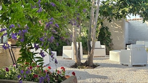 Un jardín con mobiliario exterior blanco, un árbol y plantas con flores.