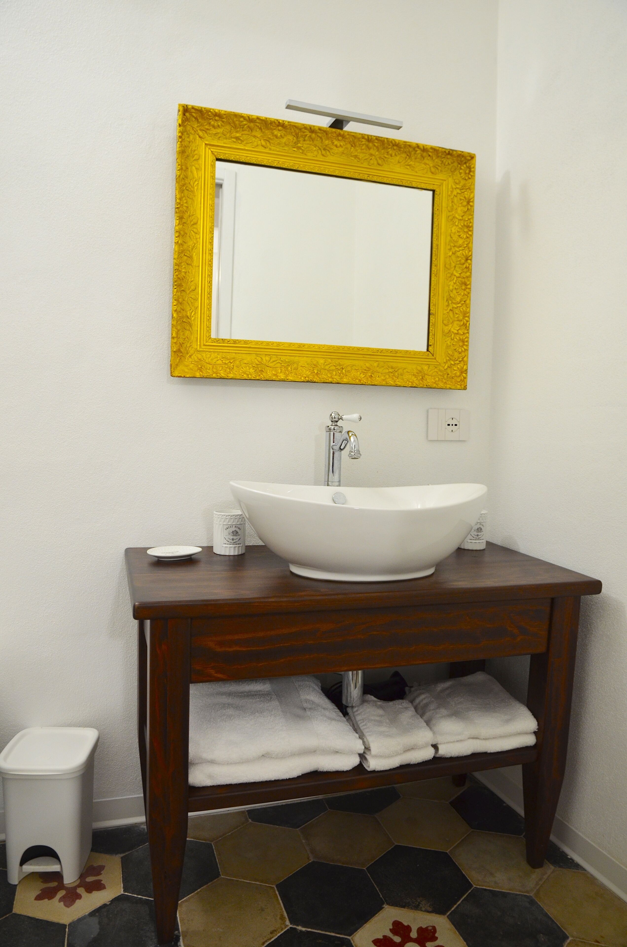 Un baño con lavamanos blanco, tocador de madera, espejo con marco amarillo y suelo de baldosas.