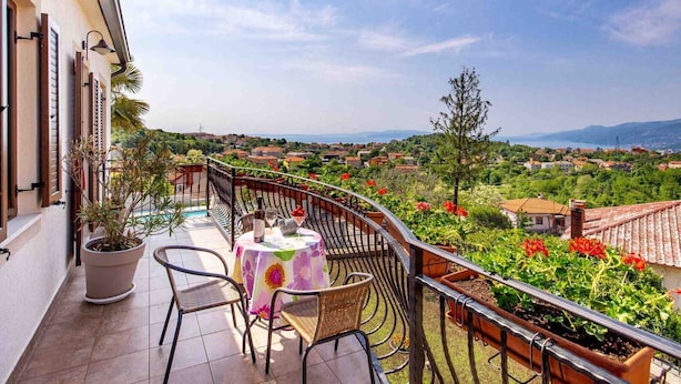 A balcony with a table and chairs, overlooking a scenic view of a town and mountains. - Modern Comfort with Shared Pool, Balconies & Lush Green Surroundings - One Bedroom Apartment (Kastav)
