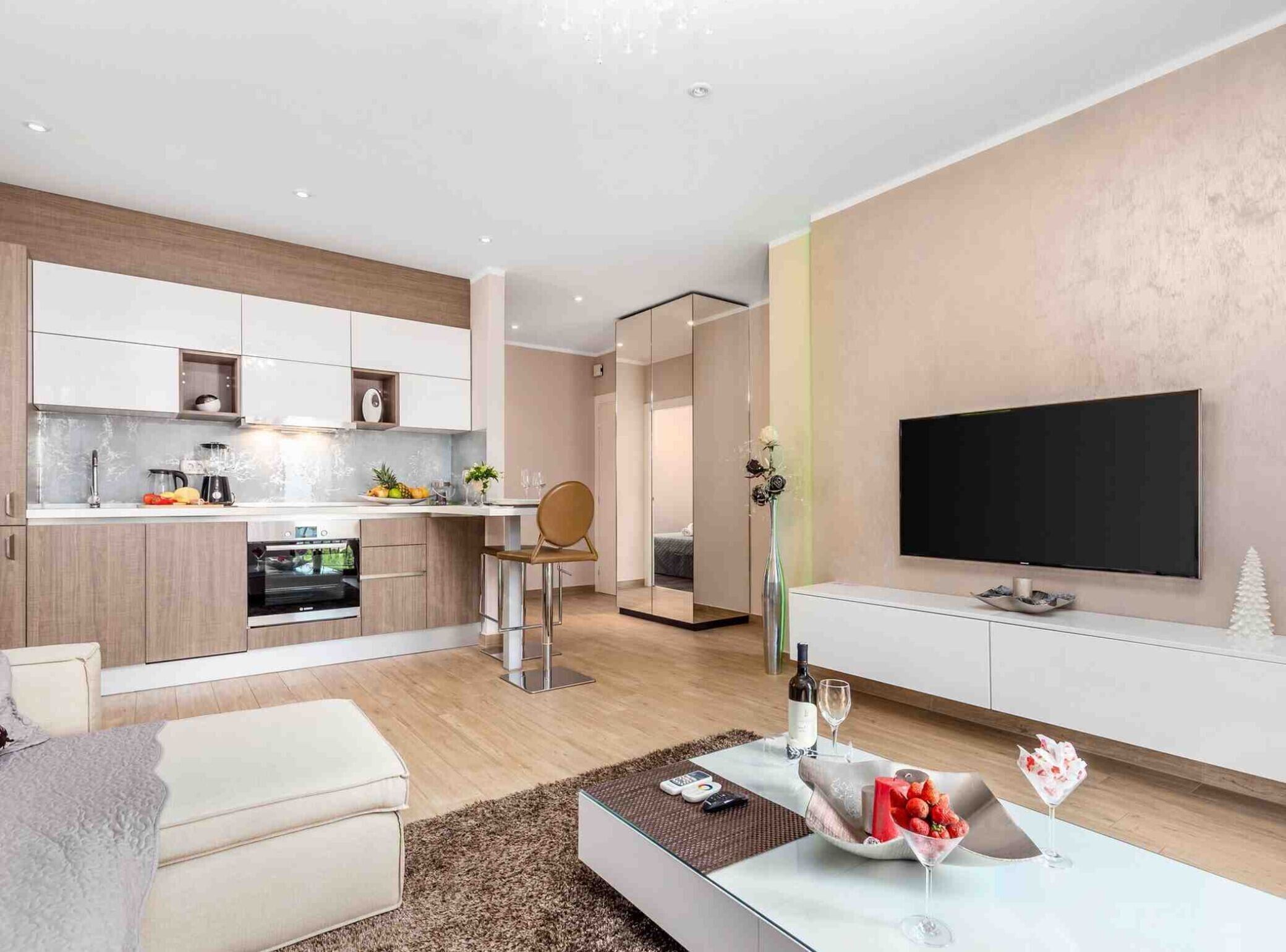 A modern living room with a sofa, a coffee table with fruit and a bottle, a TV mounted on the wall, and a kitchen area with cabinets and appliances.