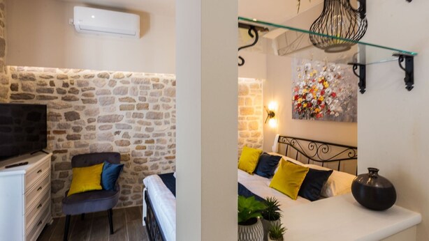 Una habitación con pared de piedra, televisor, sofá con cojines, una mesita y una estantería de pared con una planta.