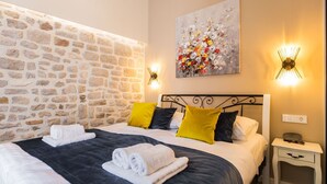 Un dormitorio con pared de piedra, cama con ropa de cama blanca y oscura, almohadas amarillas y negras, un cuadro y luces fijadas a la pared.