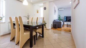 Un moderno comedor con una mesa de vidrio y sillas color beige, un área de estar con sofá y televisor, y un reloj en la pared.