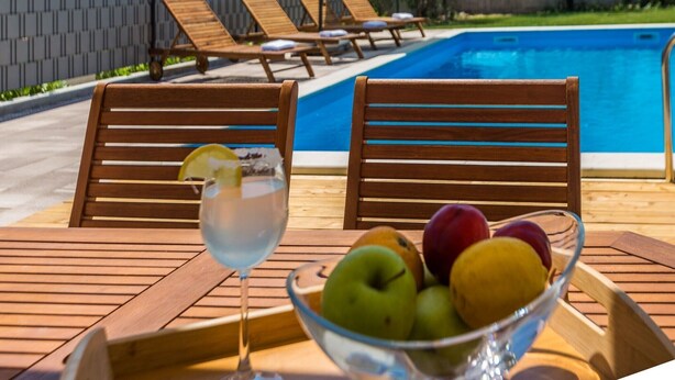 Escena junto a la piscina con sillas y una mesa de madera, un vaso de bebida y un bol de fruta.