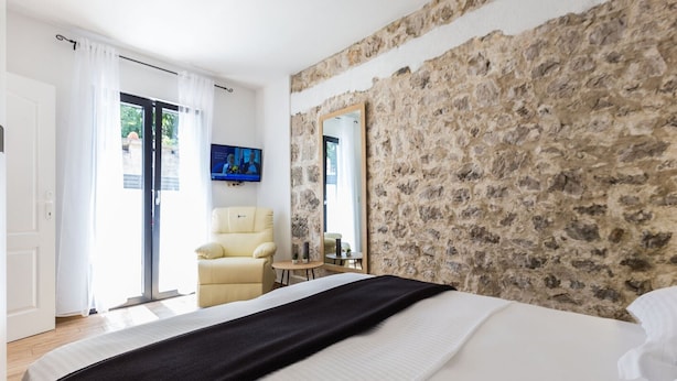 Een slaapkamer met een stenen muur, een bed met witte beddengoed, een beige fauteuil, een klein tafeltje en een televisie aan de muur.