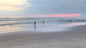 Une plage où deux personnes se tiennent dans une eau peu profonde, sous un ciel dégagé, au moment du coucher du soleil.