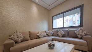 A modern living room with a beige sofa, a glass coffee table, and a large window providing a view of buildings outside.