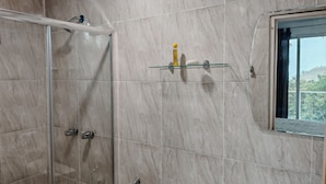 Un baño con cabina de ducha de vidrio, repisa de vidrio con artículos de aseo y ventana con vista a follaje.