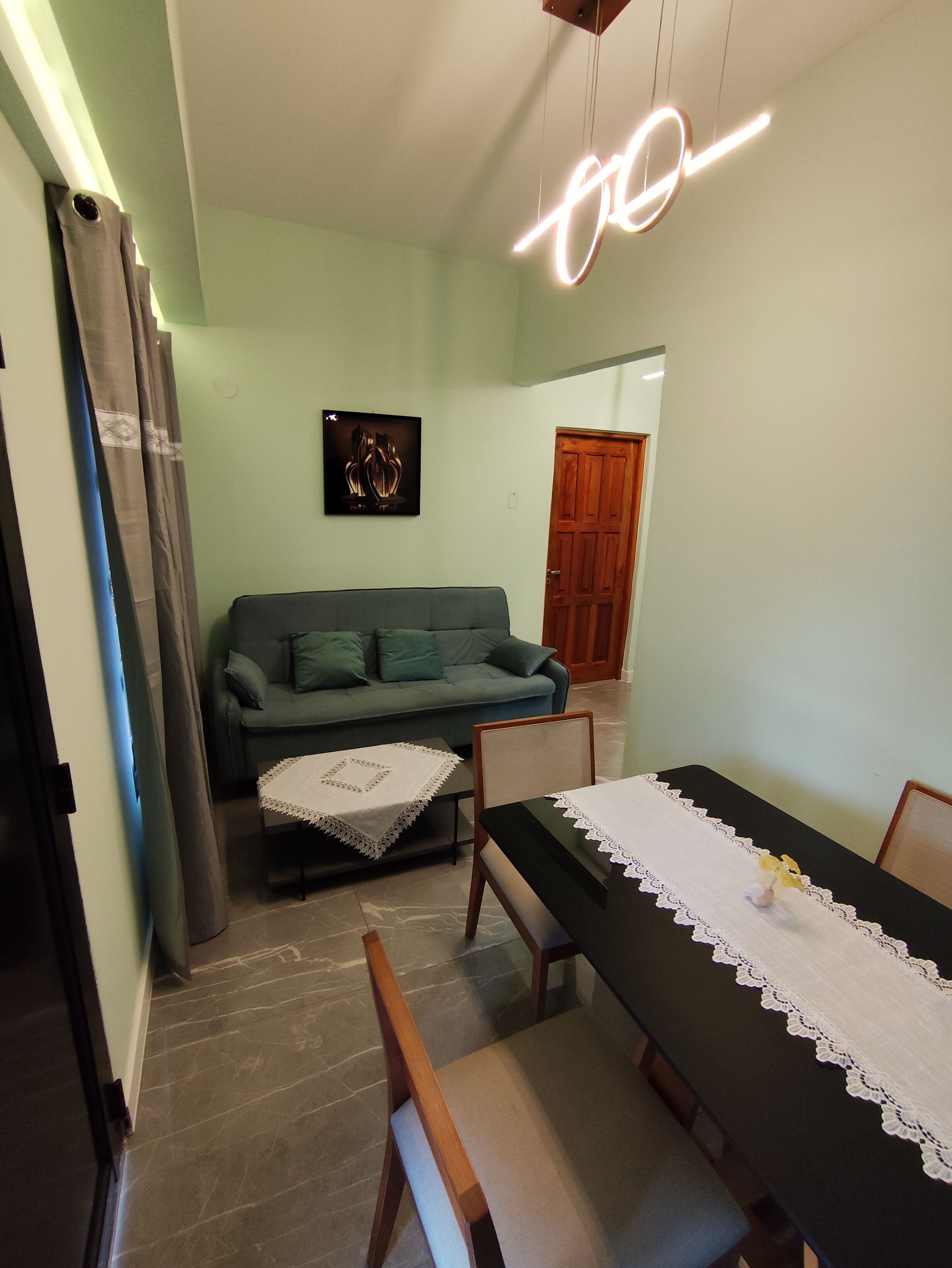 A living room with a green sofa, a small table with a white lace tablecloth, and a dining table with a black tablecloth and white lace table runner.