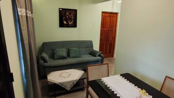 A living room with a green sofa, a small table with a white lace tablecloth, and a dining table with a black tablecloth and white lace table runner.