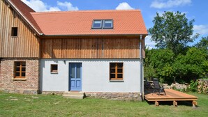 Une maison à deux étages, avec un toit recouvert de tuiles rouges, une porte bleue et une petite terrasse aménagée avec du mobilier d’extérieur.