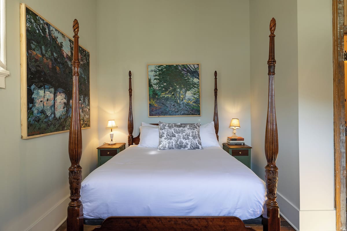 A four-poster bed with white linens, two nightstands with lamps, and two paintings on the walls.