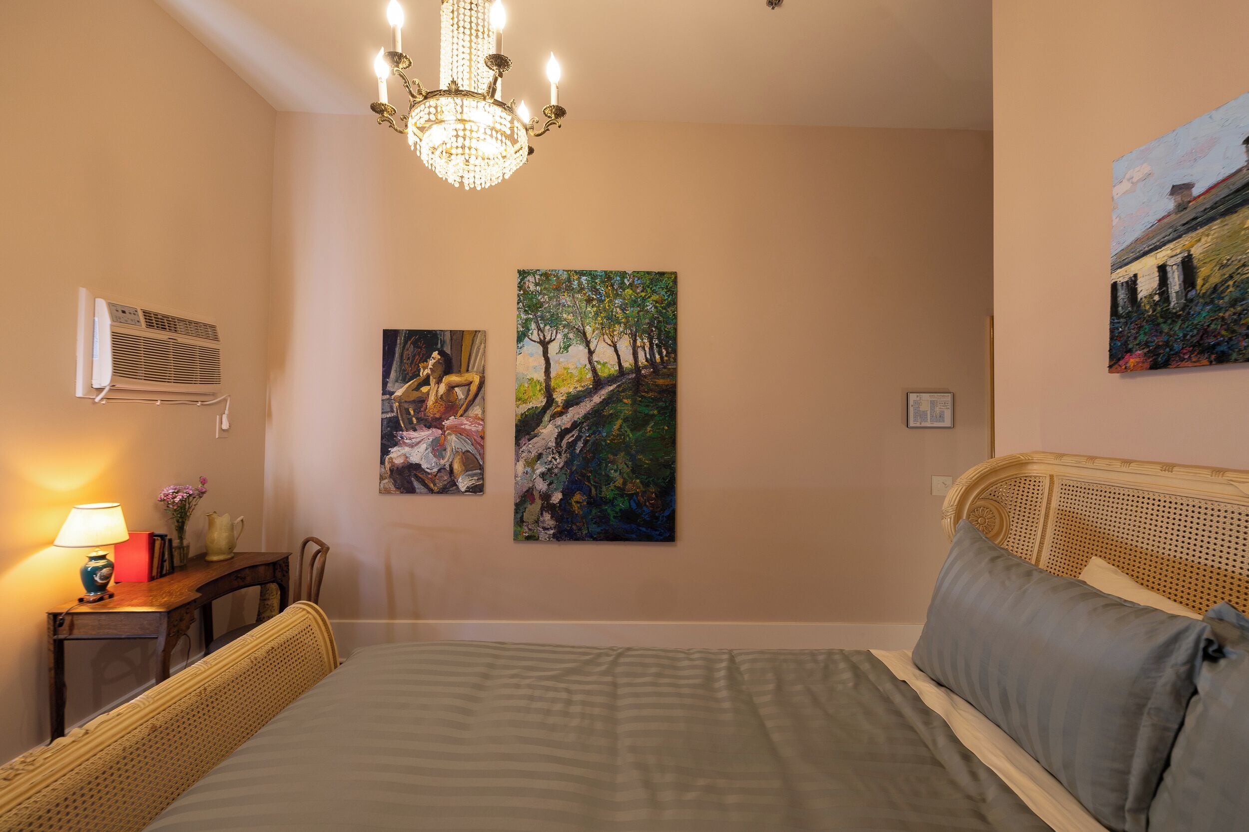 A bedroom with a bed, a chandelier, an air conditioner, and two paintings on the wall.