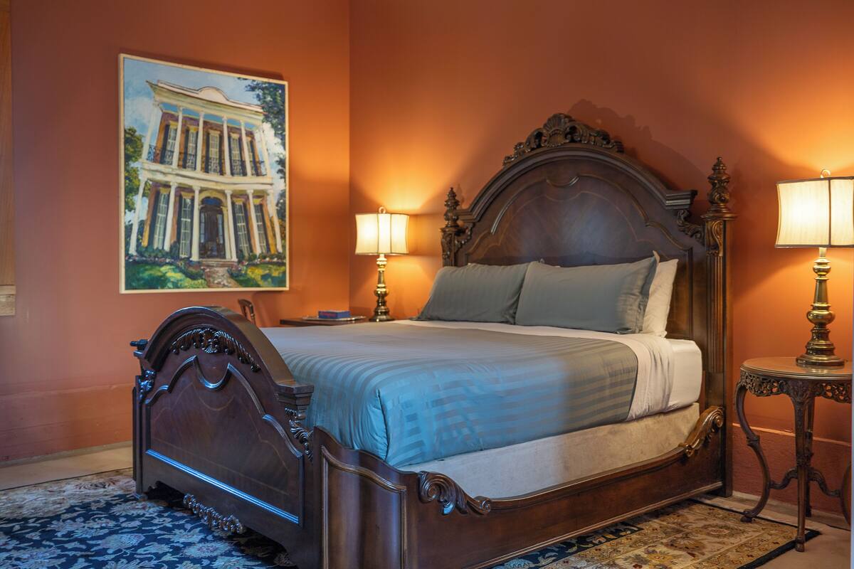 A large, ornate bed with a wooden headboard, two bedside lamps, and a large painting of a building on the wall.