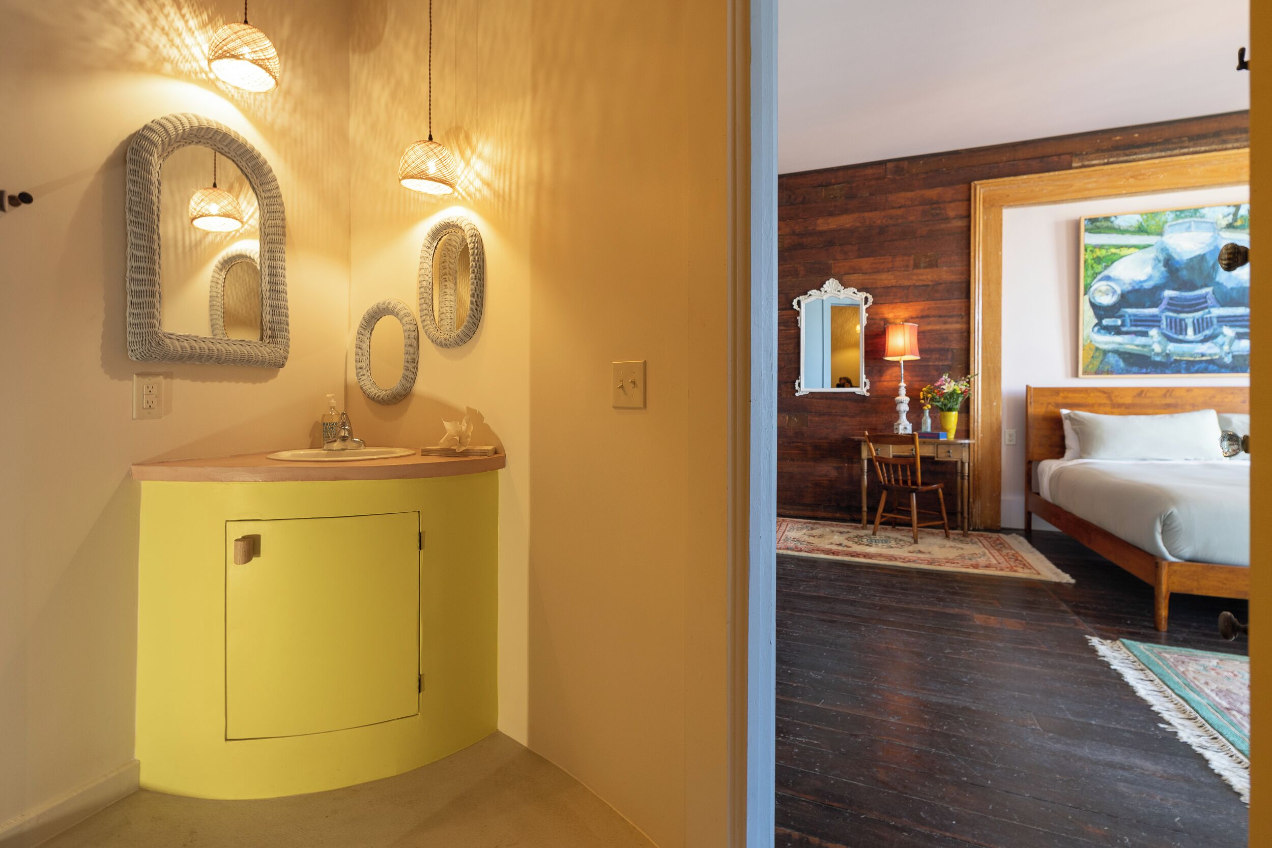 Ein gelbes Badezimmer mit Waschbecken und Spiegeln, ein Holztisch mit Lampe sowie ein Bett mit einem Gemälde.