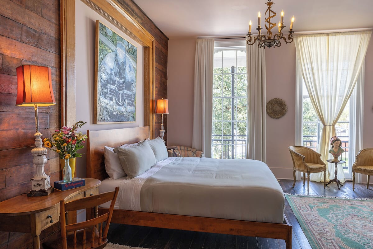A bedroom with a large bed, wooden headboard, two bedside lamps, a chandelier, a window with curtains, and a small table with chairs.