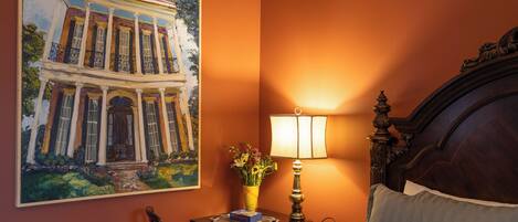 Habitación de hotel con un cuadro grande de un edificio de estilo colonial, una lámpara de noche, un jarrón con flores y una cama bien hecha.