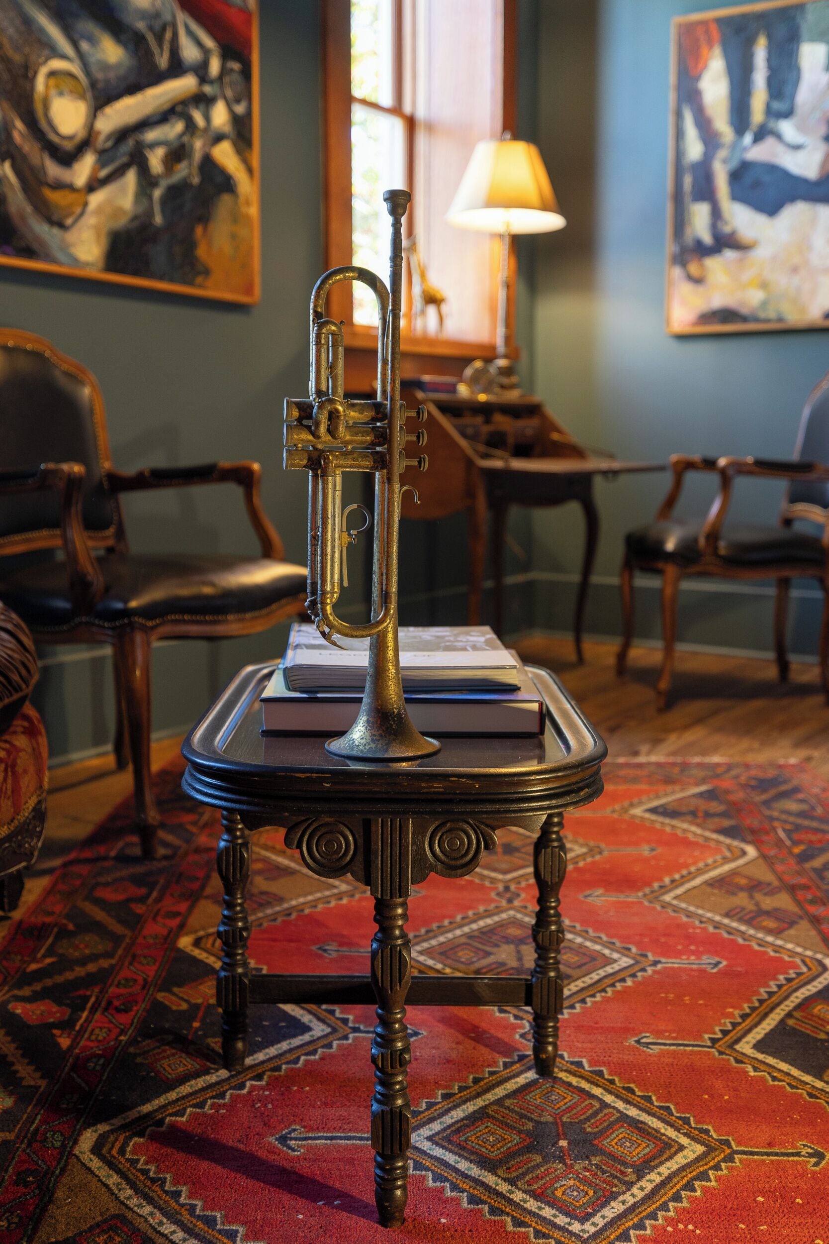A room with a vintage brass trumpet on a table, surrounded by chairs and a patterned rug.