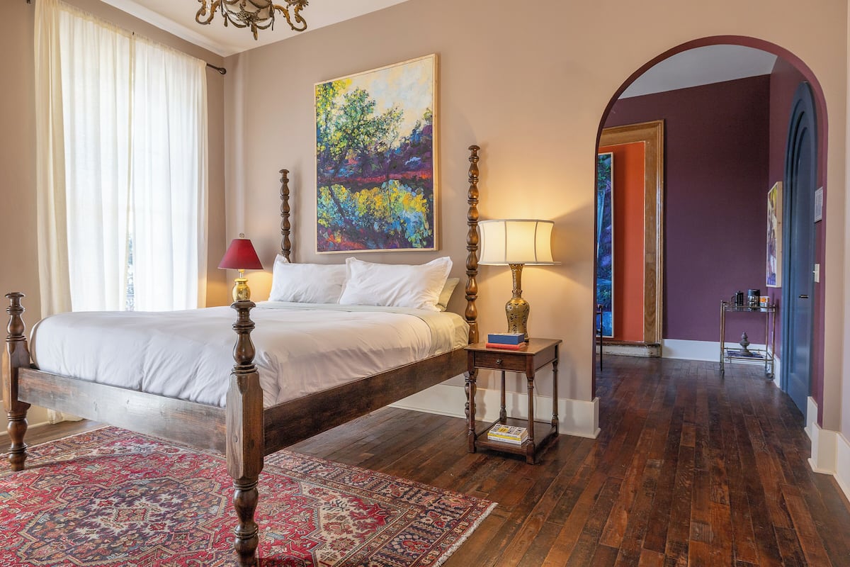 A bedroom with a four-poster bed, a red lamp, a painting, and a wooden floor.