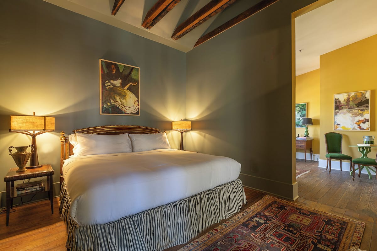 A bedroom with a large bed, two bedside lamps, a painting on the wall, and a view into another room.