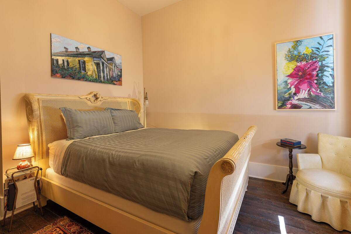 A neatly arranged bedroom with a bed, a chair, a small table, and two paintings on the wall.