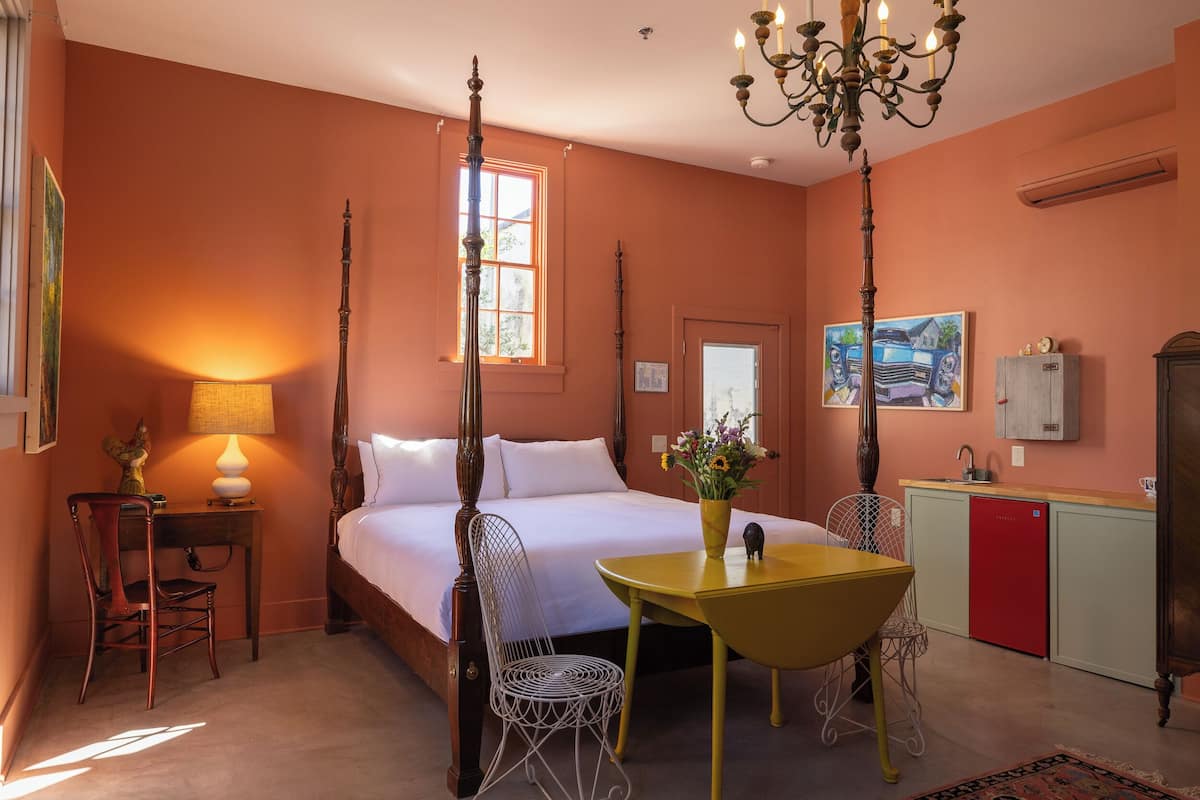 A room with a four-poster bed, a dining table with chairs, a chandelier, a desk with a chair, a red refrigerator, and a painting on the wall.