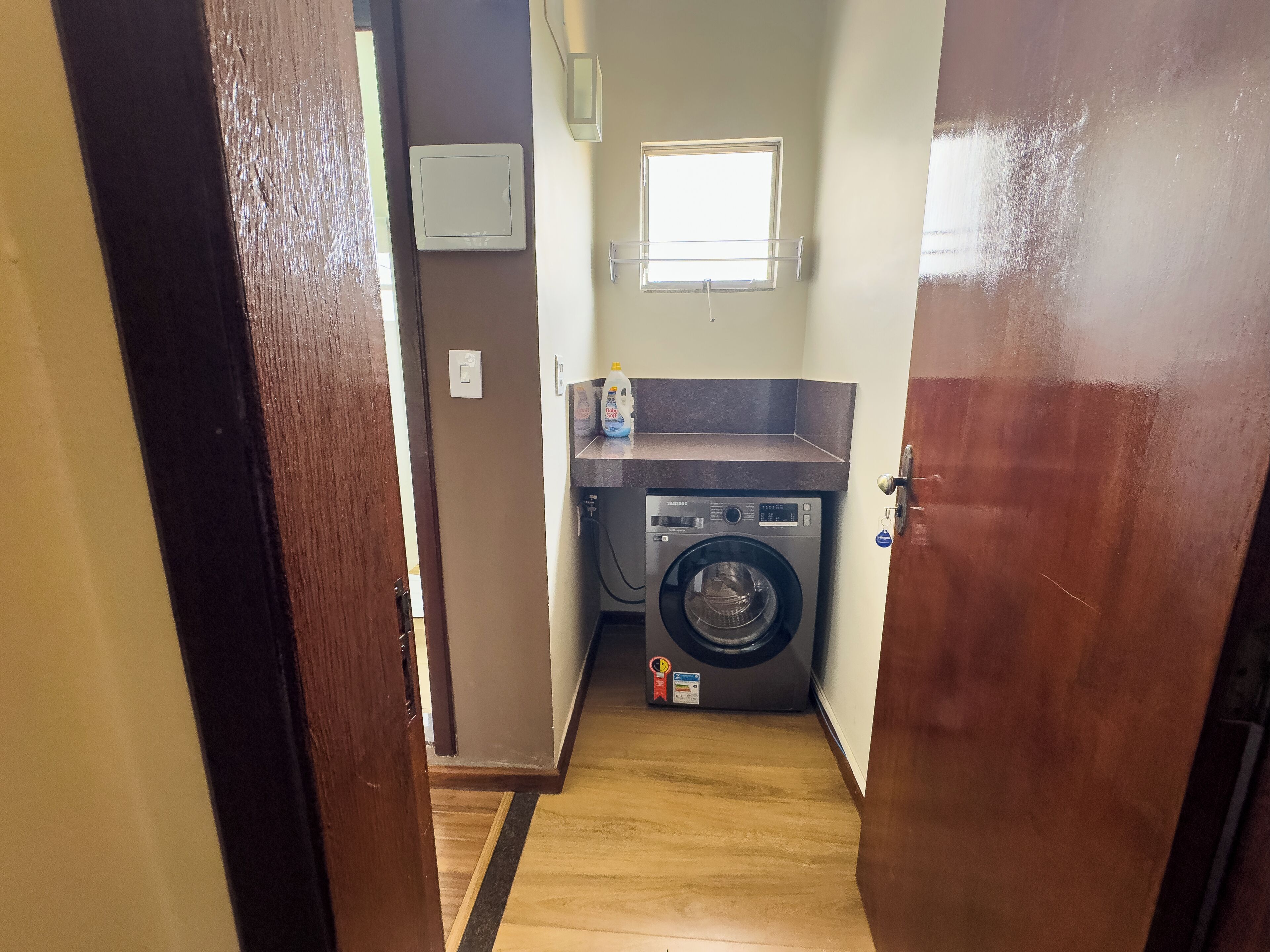 A laundry room with a washing machine, a small window, and a countertop with a bottle of detergent.