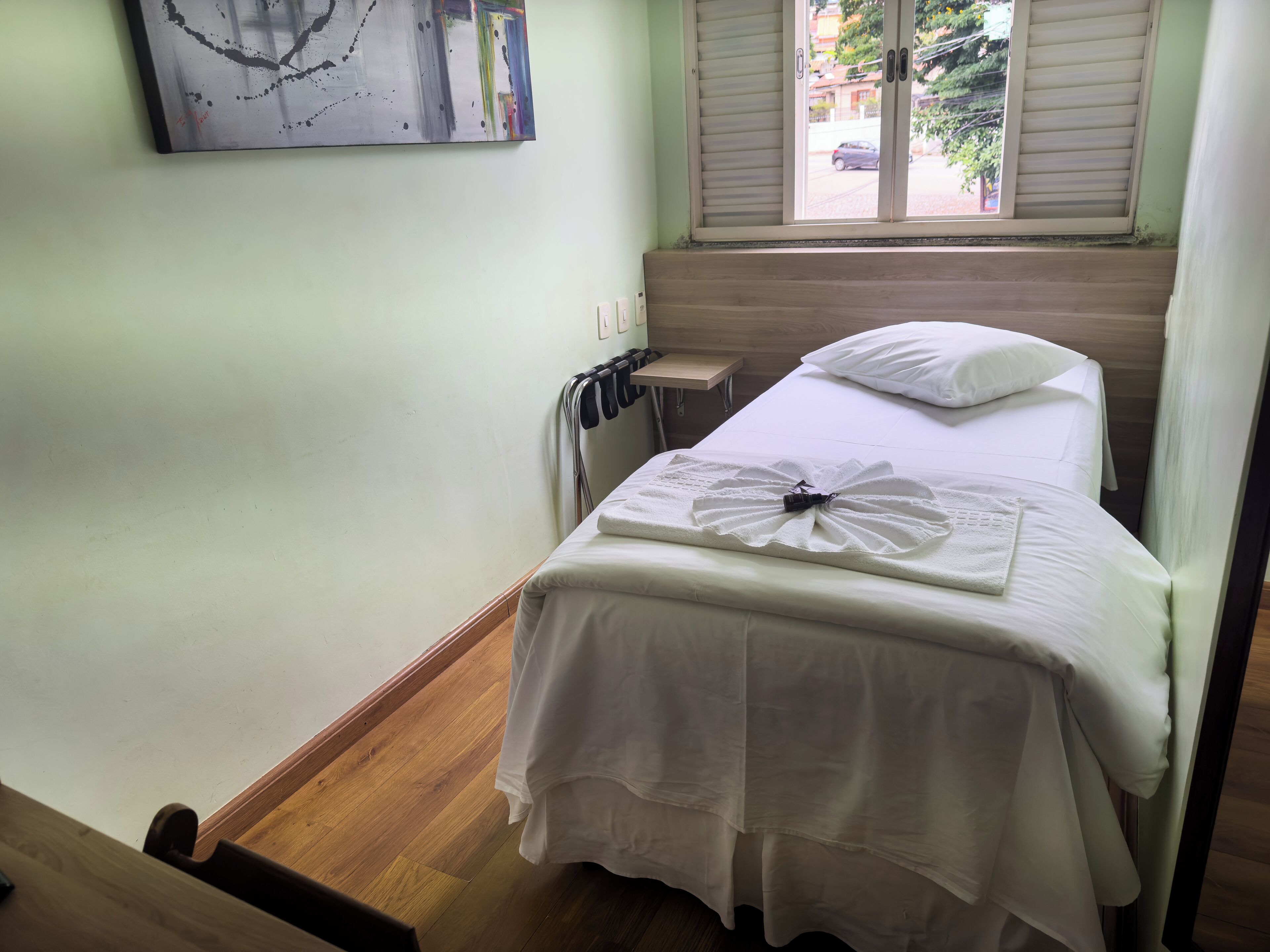 Een hotelkamer met een bed, een klein nachtkastje, een kunstwerk aan de muur en een raam met zonwering.