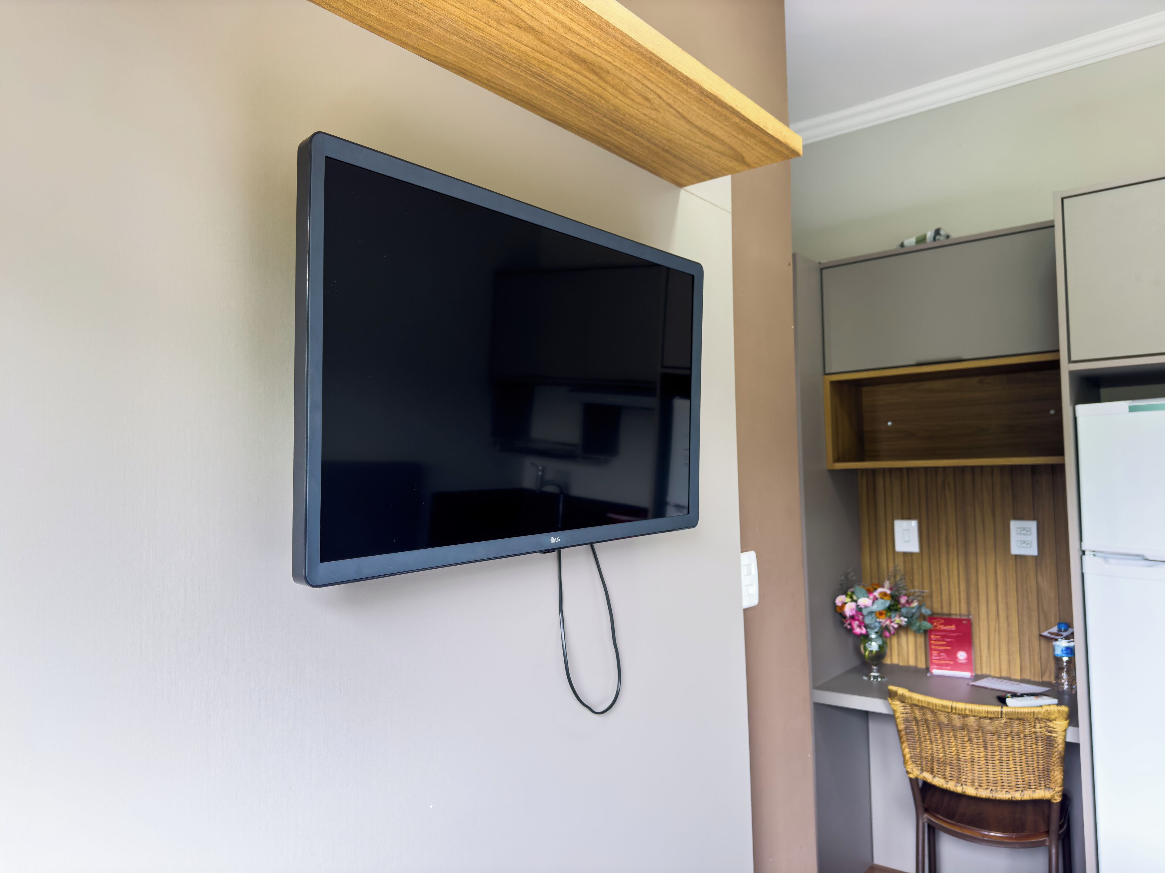 Un televisor de pantalla plana montado en la pared en una habitación con techo de madera, una kitchenette con refrigerador y una silla de mimbre.
