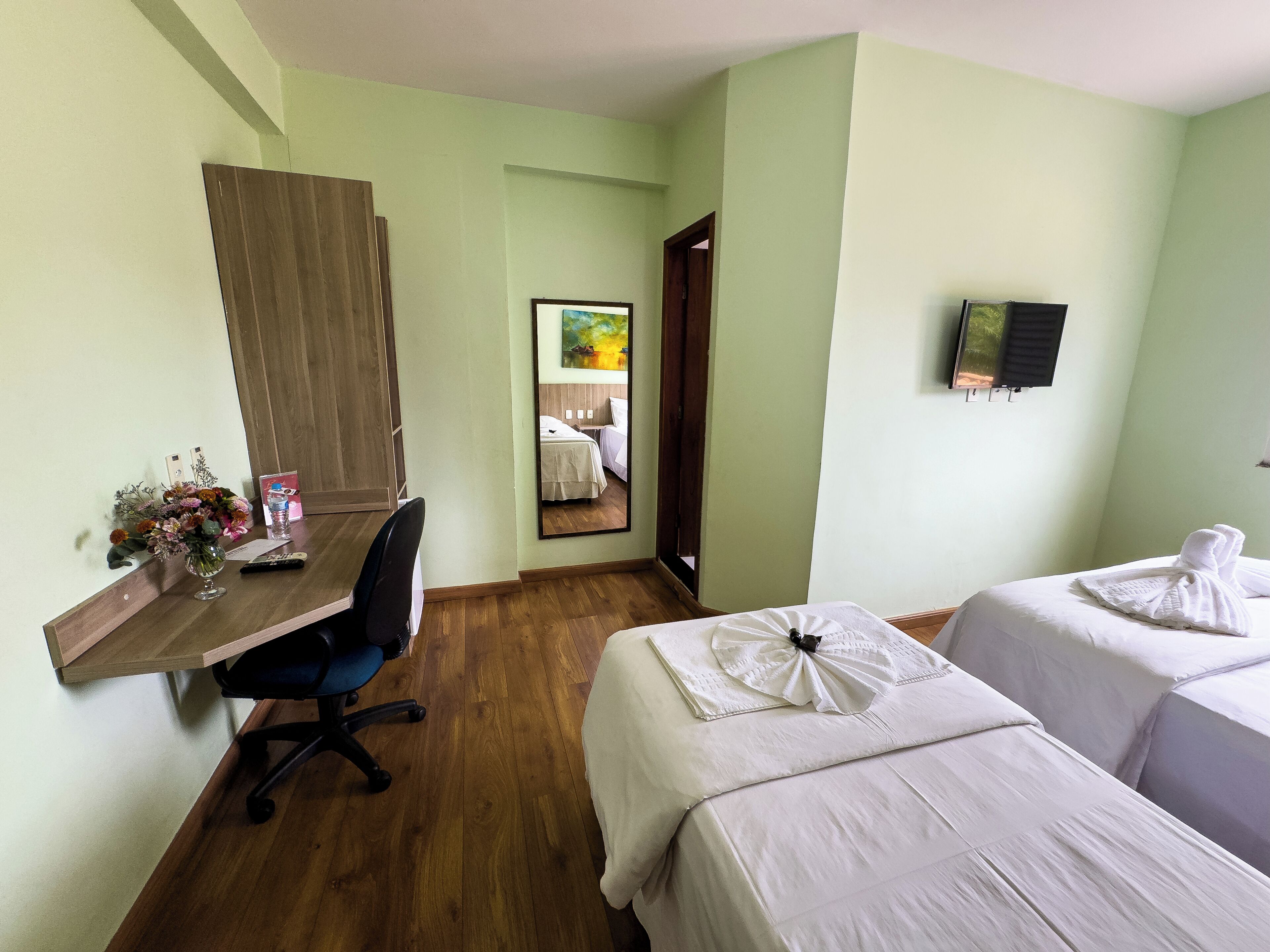 A hotel room with a wooden desk, a chair, a bed with white linens, a small table with a vase of flowers, and a mirror on the wall.