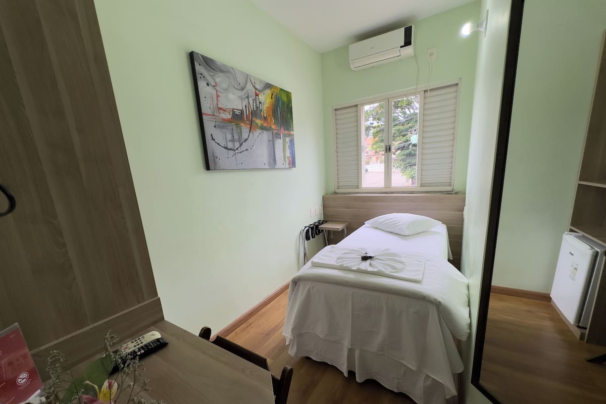 Une chambre d’hôtel avec un lit, une petite table de chevet, un miroir, une fenêtre avec des stores, un appareil de climatisation et un tableau au mur.