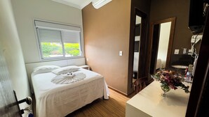 Hotellihuone, jossa on sänky, ikkuna, jossa on sälekaihtimet, puulattia, kylpyhuone, jossa on pesuallas ja peili, sekä maljakko kukkineen tasolla.