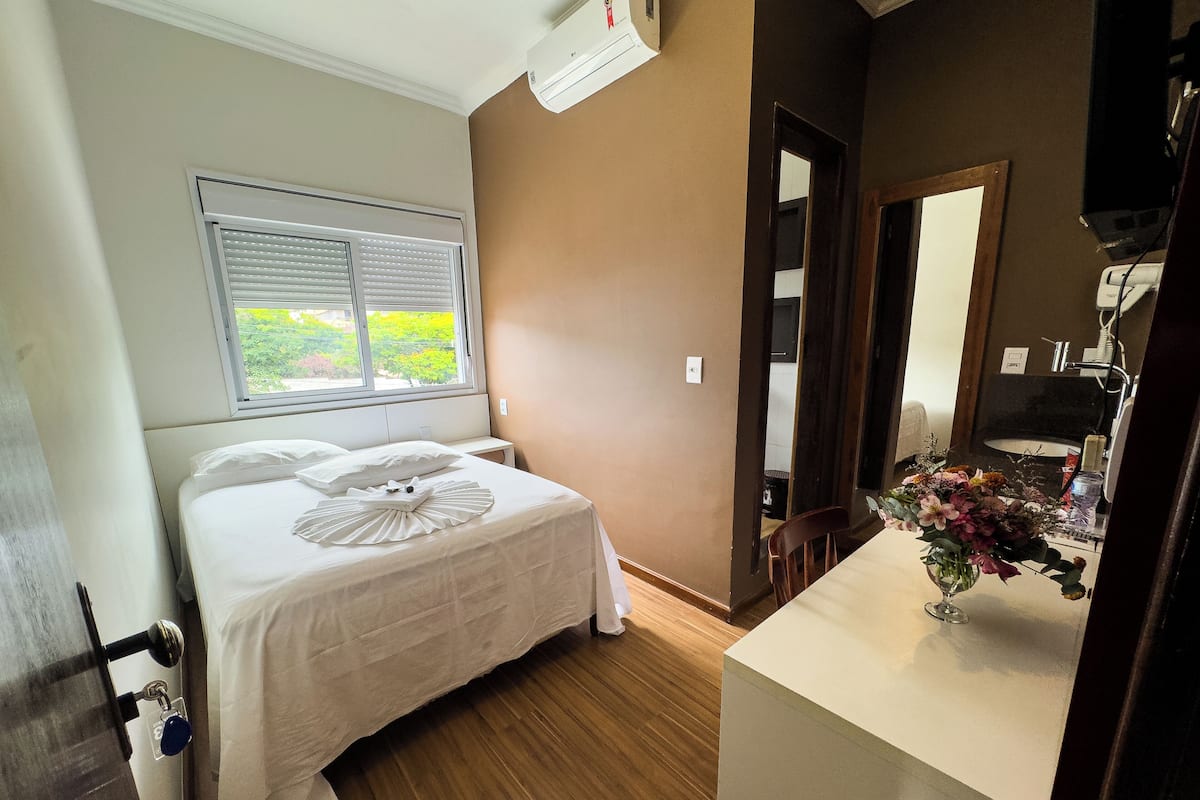 Une chambre d’hôtel avec un lit, une fenêtre avec des stores, un plancher en bois, une salle de bain avec un lavabo et un miroir, et un vase avec des fleurs posé sur le comptoir.