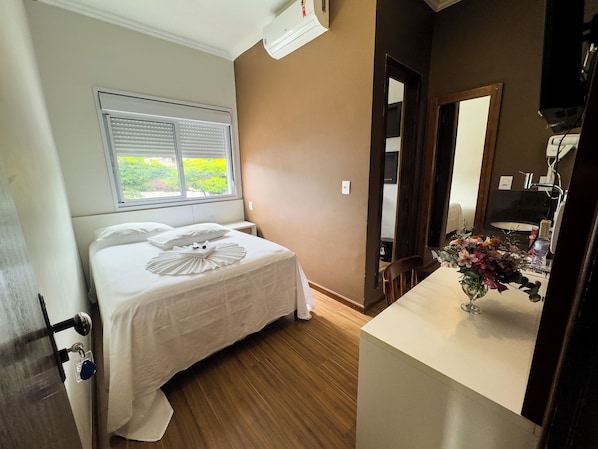 A hotel room with a bed, a window with blinds, a wooden floor, a bathroom with a sink and mirror, and a vase with flowers on the counter.