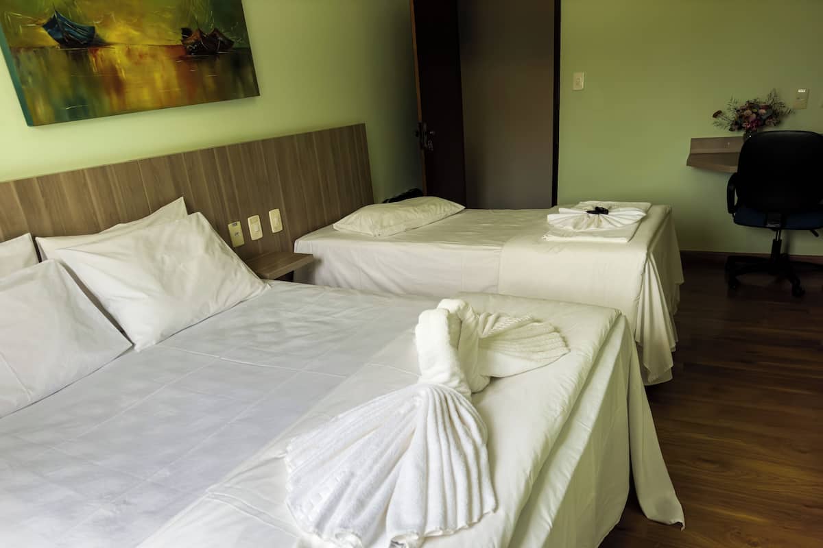 Une chambre d’hôtel avec deux lits, chacun agrémenté de draps et de serviettes blancs. On y trouve une tête de lit en bois, une petite table et une chaise. Un tableau est accroché au mur, et un vase rempli de fleurs est posé sur une étagère.