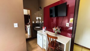 Kompakti hotellihuone, jossa on valkoinen työpöytä, tuoli, pieni valkoinen kaappi, peili, punaisella värillä korostettu seinä, seinälle asennettu televisio ja puulattia.