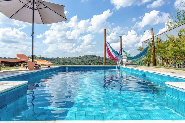 A clear blue pool with a hammock and lounge chairs, surrounded by greenery and a view of hills. - Casa com piscina - Brazilian Corner (Ibiuna)